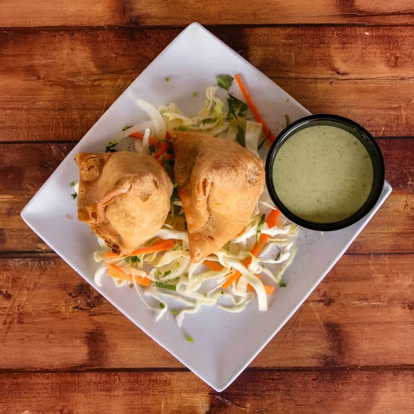 Two golden-brown samosas on a bed of shredded cabbage and carrots, with a side of green chutney.