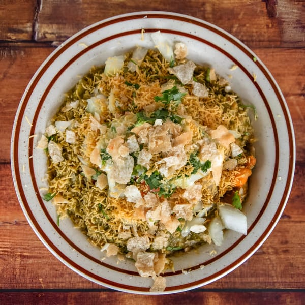 Overhead view of a bowl of Indian chaat, topped with crispy noodles, chopped onions, and cilantro.