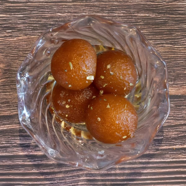 Four glistening gulab jamuns, an Indian dessert, in a clear glass bowl on a wooden table.